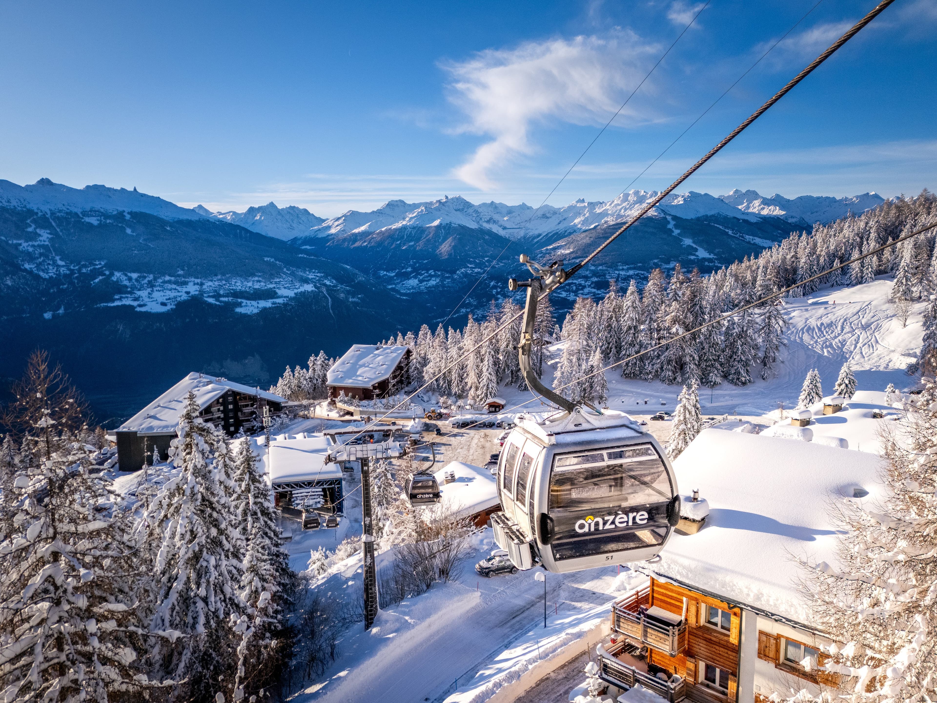 Télécabine marquée « anzère » au-dessus des chalets enneigés, sapins couverts de neige et Alpes valaisannes au fond