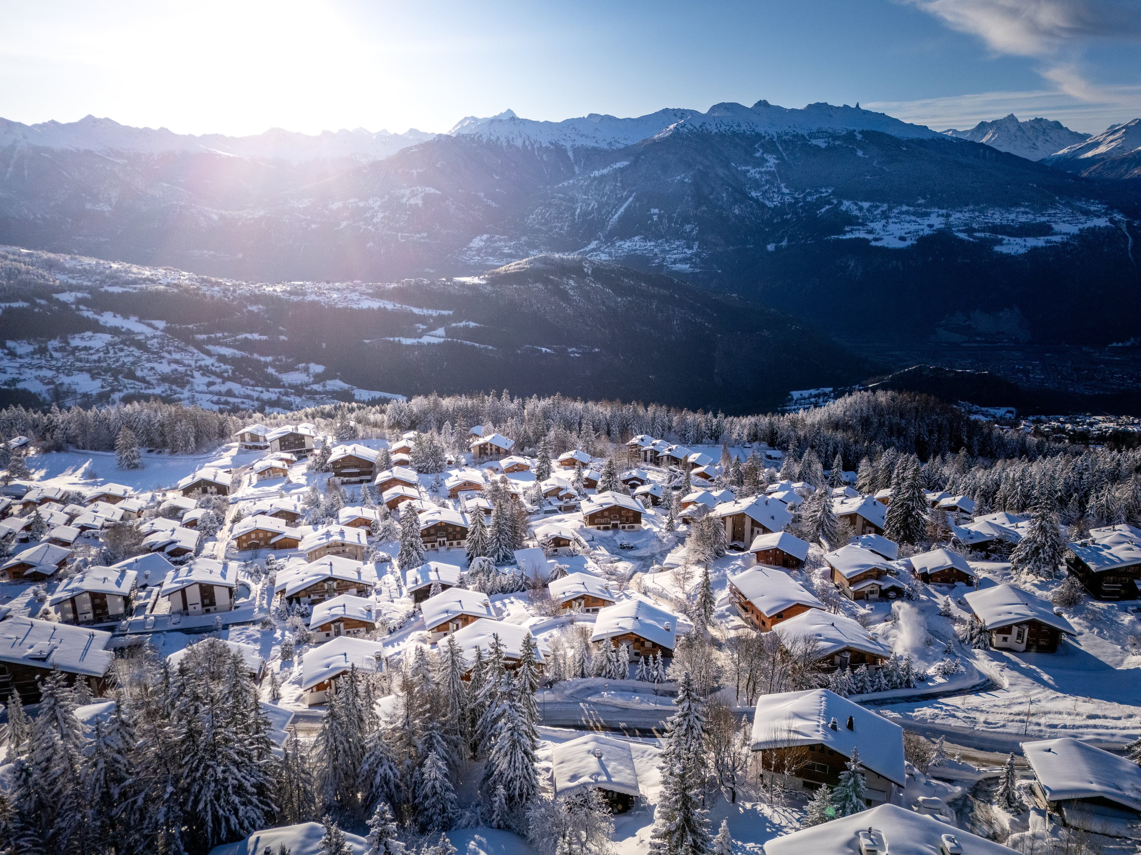 Vue aérienne d’Anzère sous la neige fraîche, chalets et forêt, la vallée du Rhône et les Alpes à contre-jour