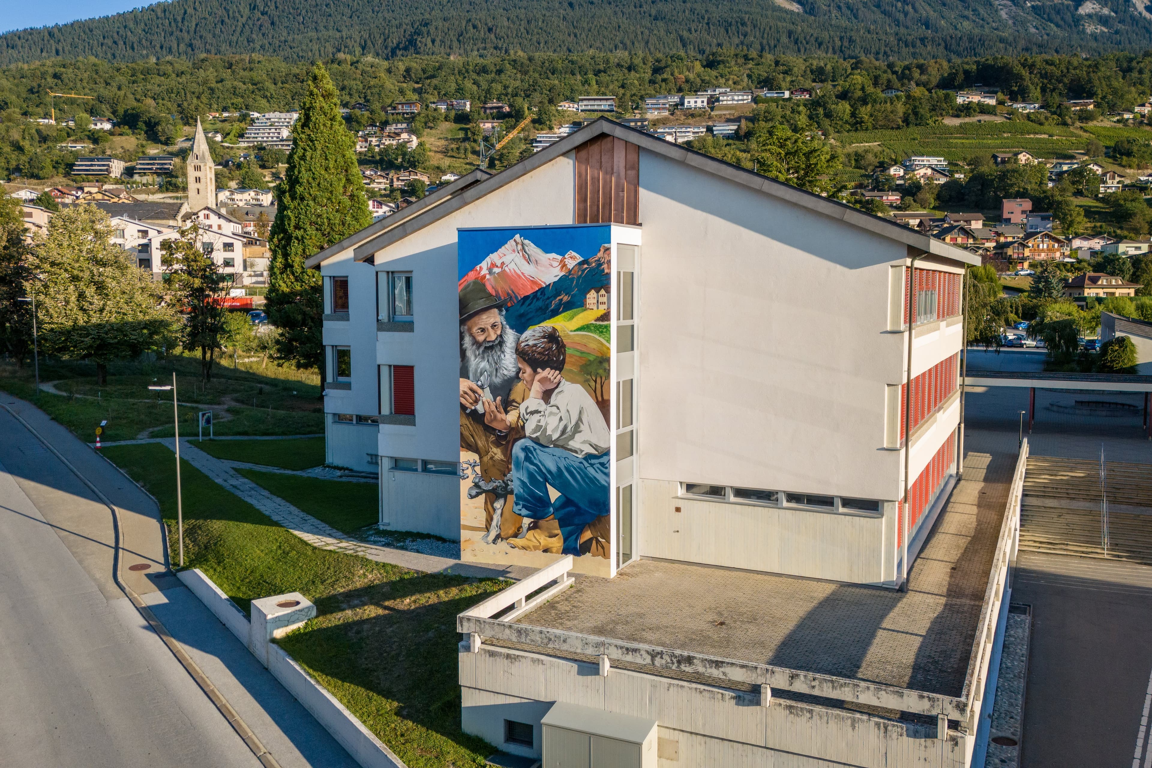 Bâtiment d’école orné d’une grande fresque, un village et son clocher derrière