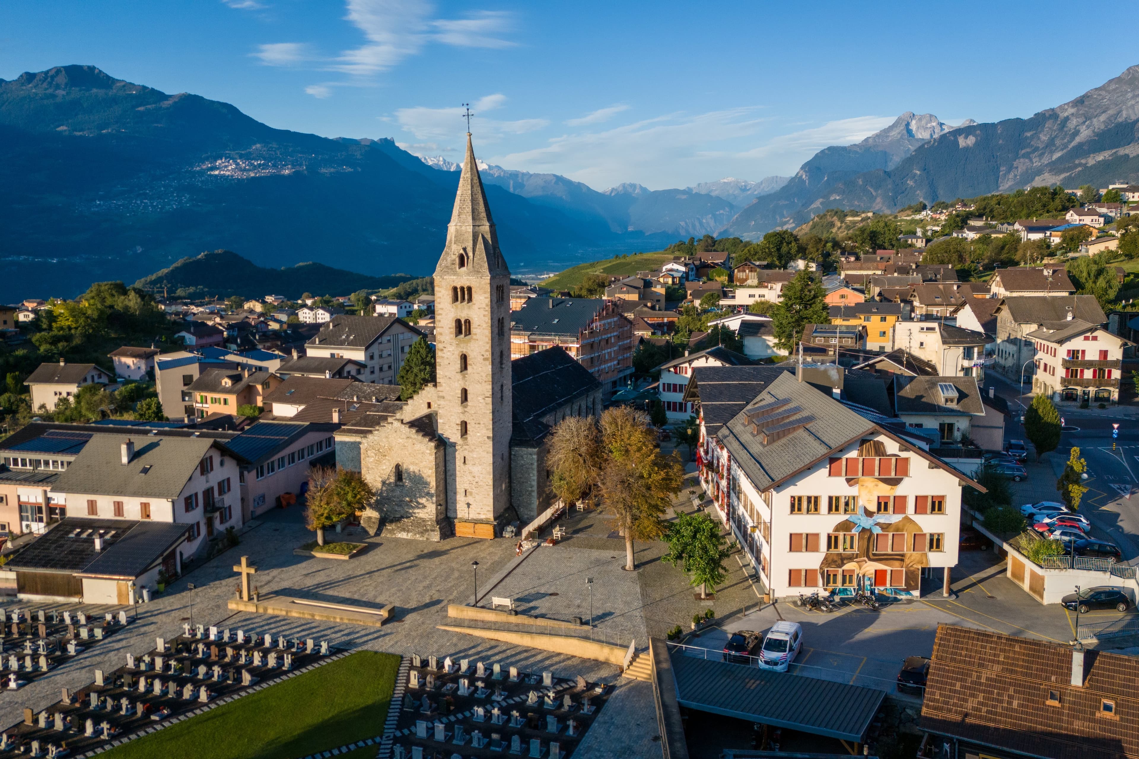 Vue au drone d’un village de Savièse : l’église au clocher pointu, la place et les maisons, montagnes au fond