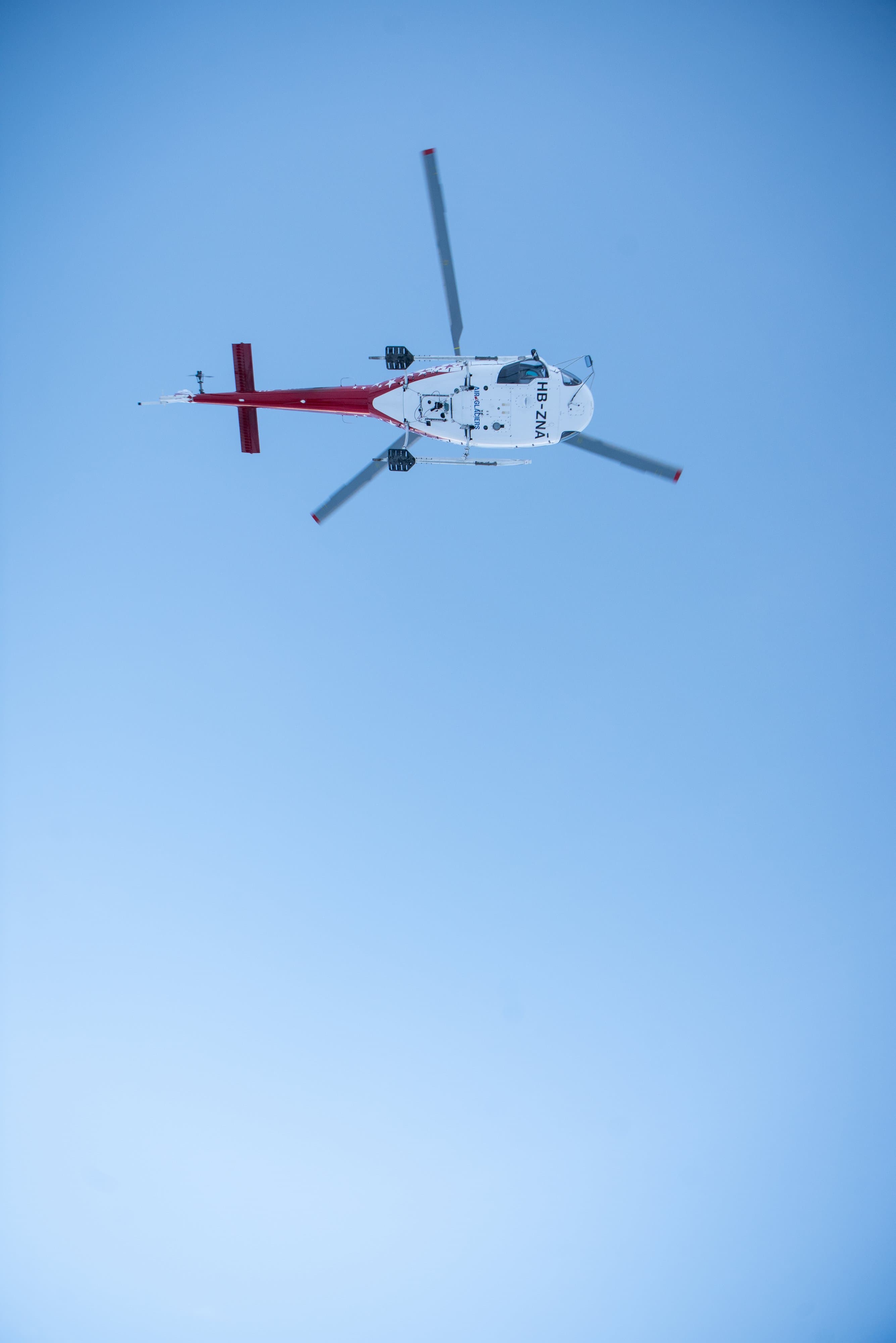 Hélicoptère rouge et blanc en vol, vu d’en dessous dans un ciel bleu