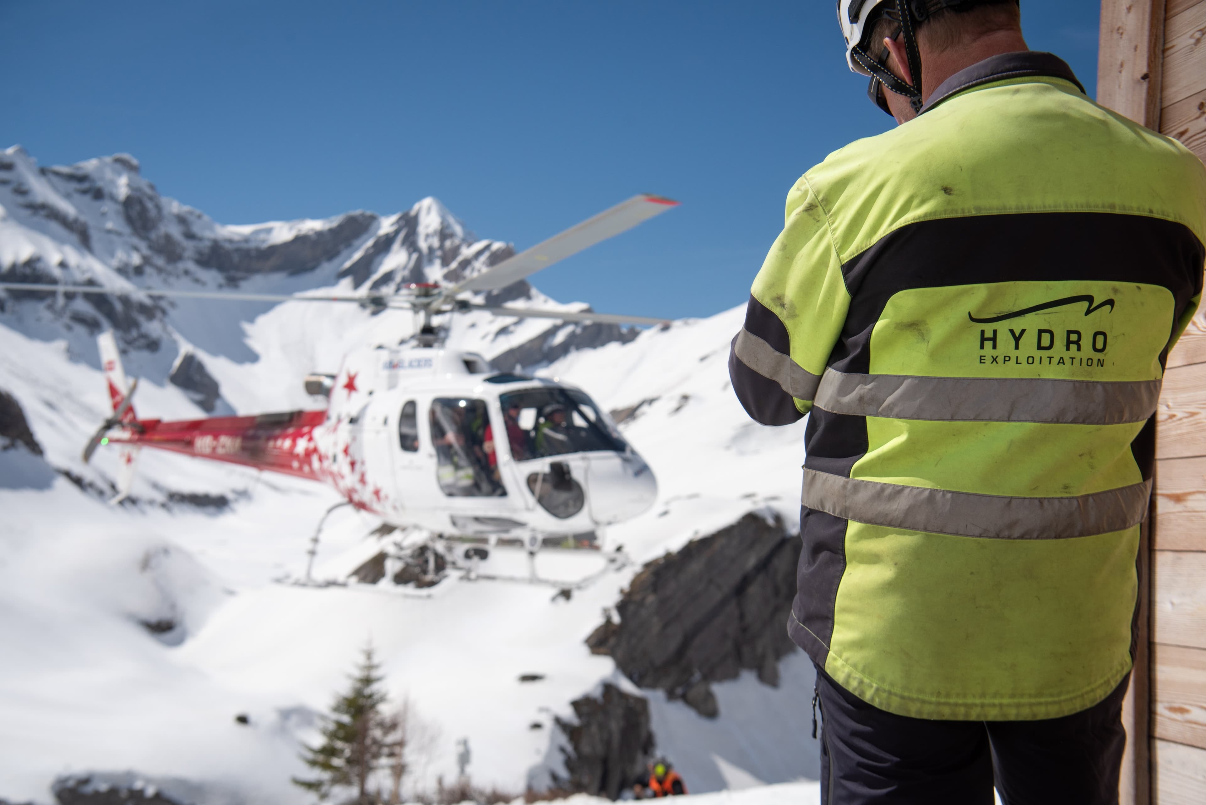 Technicien de dos en veste jaune Hydro Exploitation, un hélicoptère posé dans la neige face aux sommets