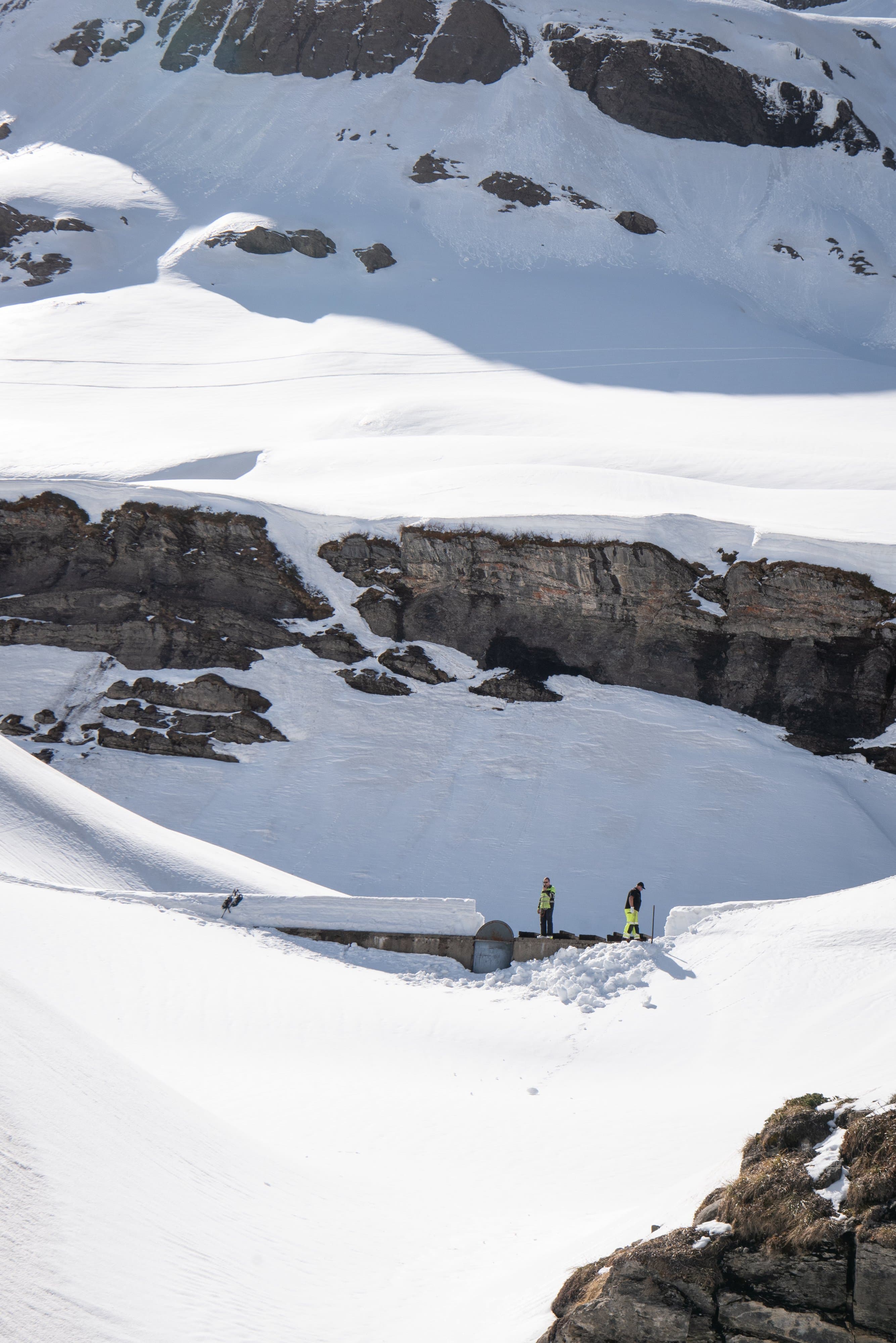 Vallon enneigé entouré de barres rocheuses, quelques techniciens près d’un chalet presque enseveli