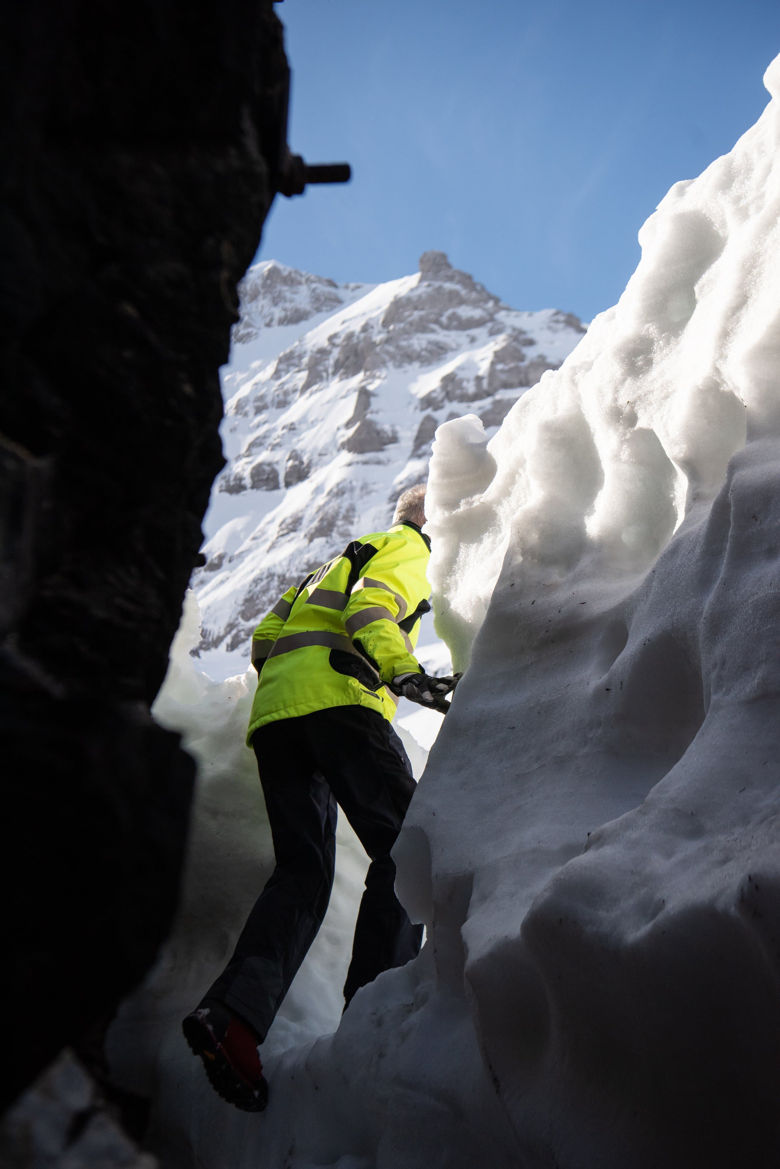 Technicien en veste jaune qui gravit un mur de neige à la sortie d’une galerie, sommets au fond