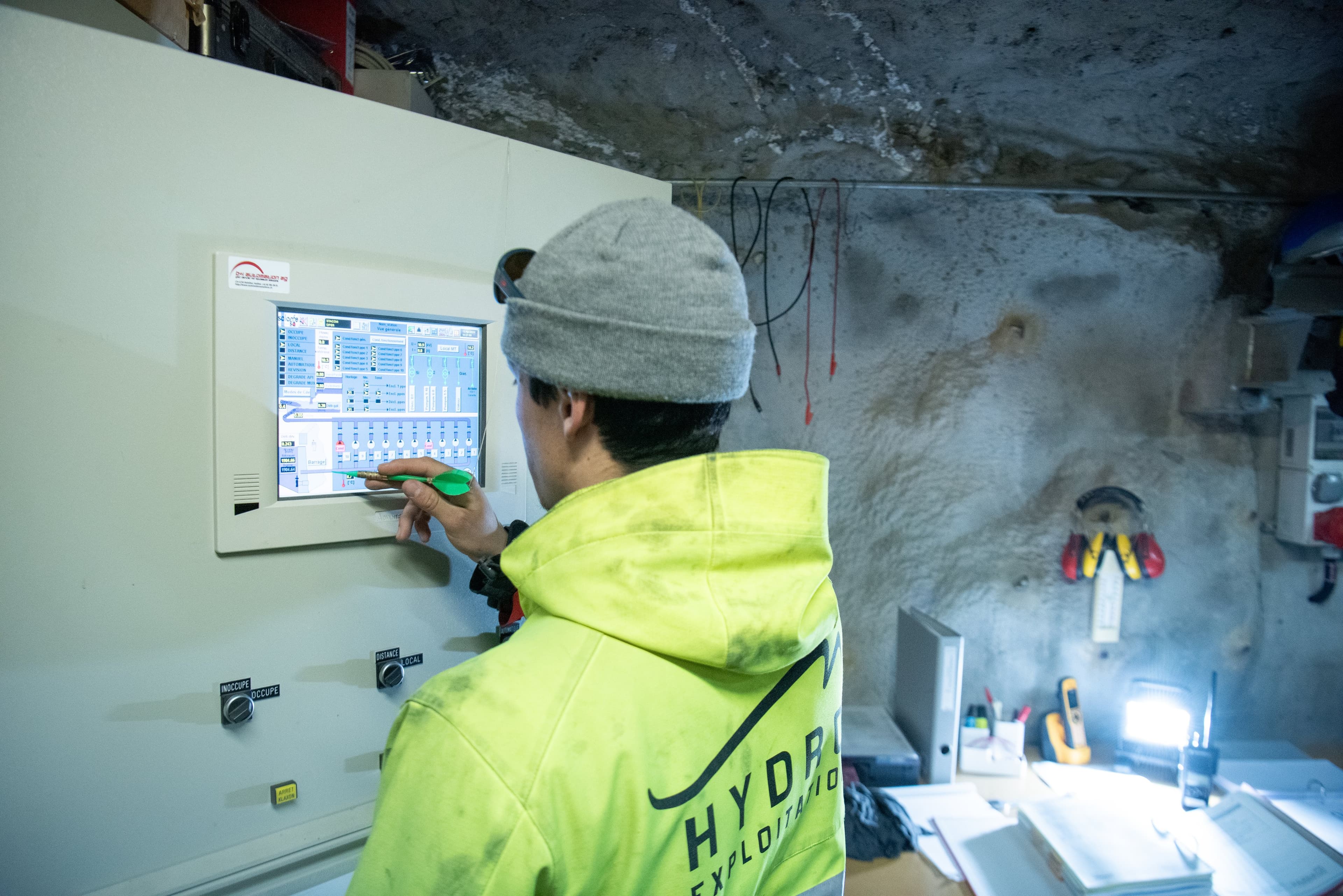 Technicien en bonnet et veste Hydro Exploitation qui règle une installation sur un écran tactile, dans une salle creusée dans la roche