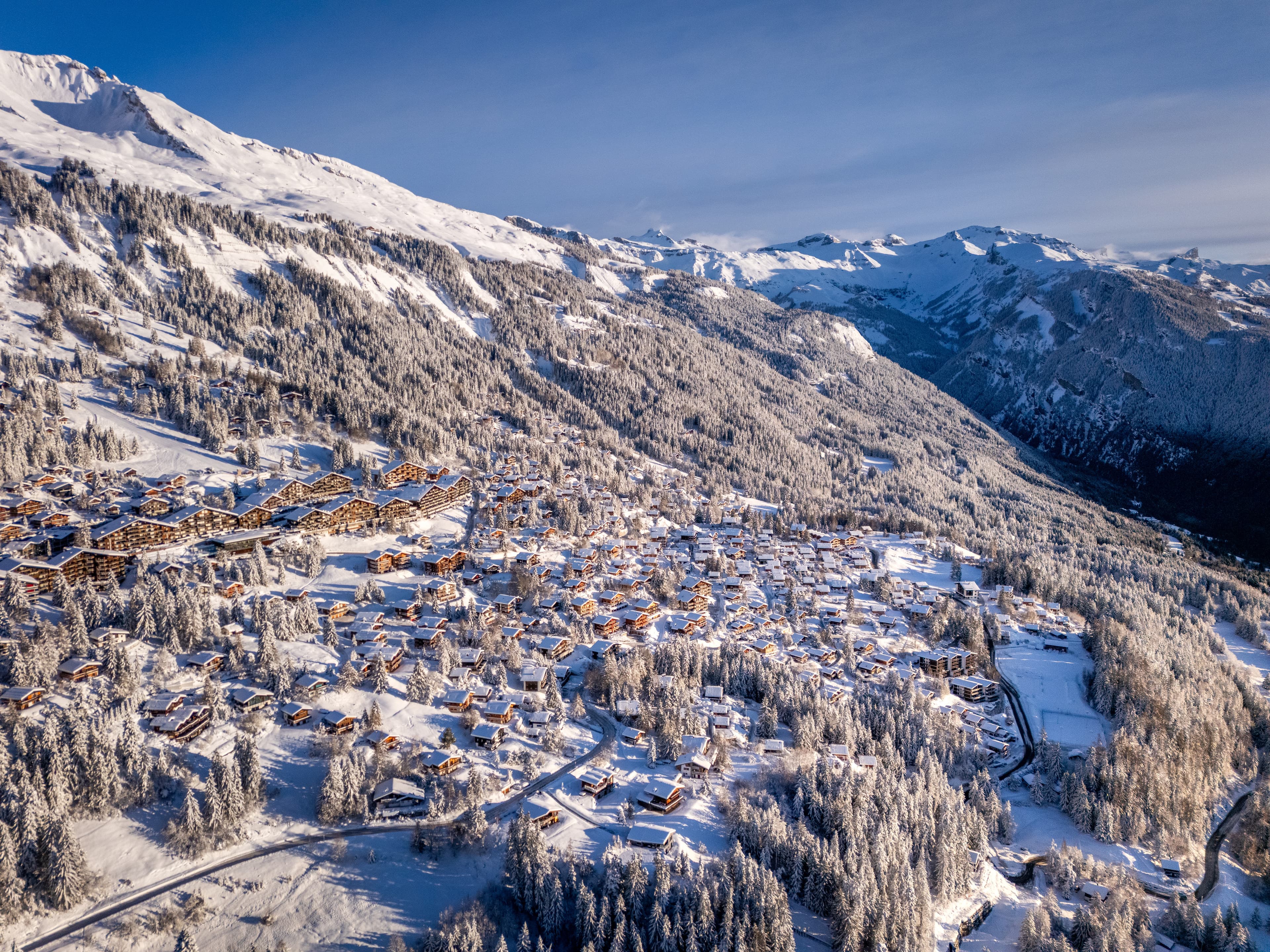 Vue aérienne d’Anzère en hiver : chalets enneigés dans la forêt, sommets au loin