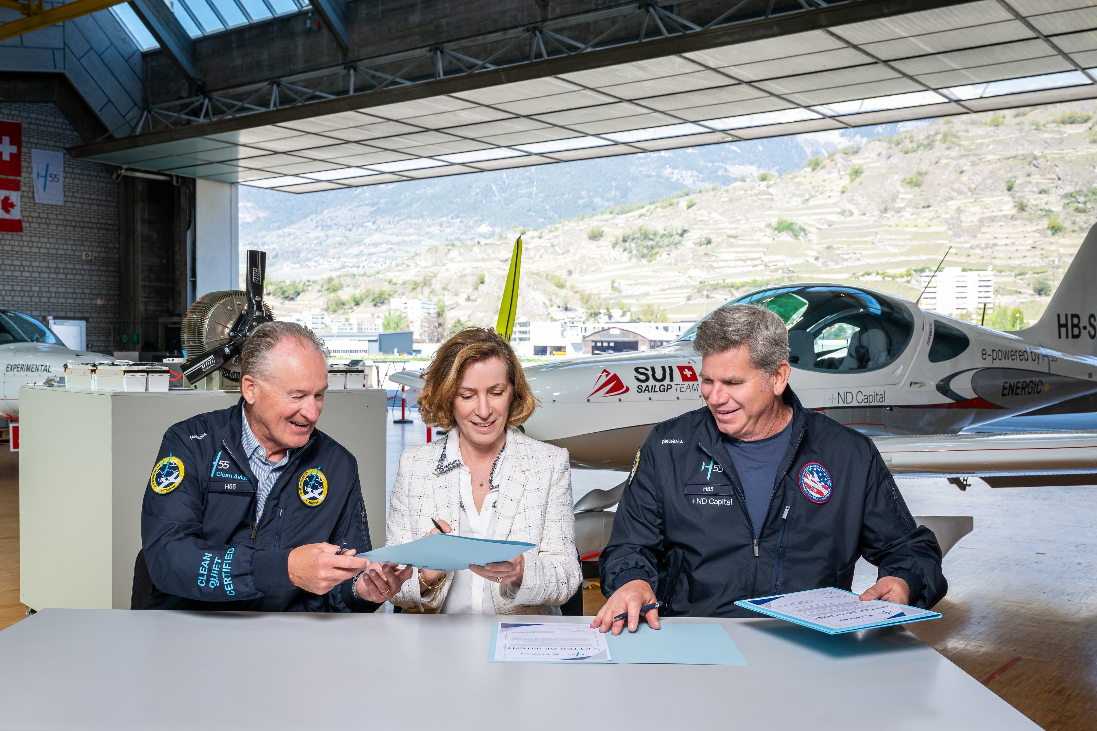 Trois signataires assis à une table dans le hangar H55, devant un avion électrique et les vignes de Sion par la porte ouverte