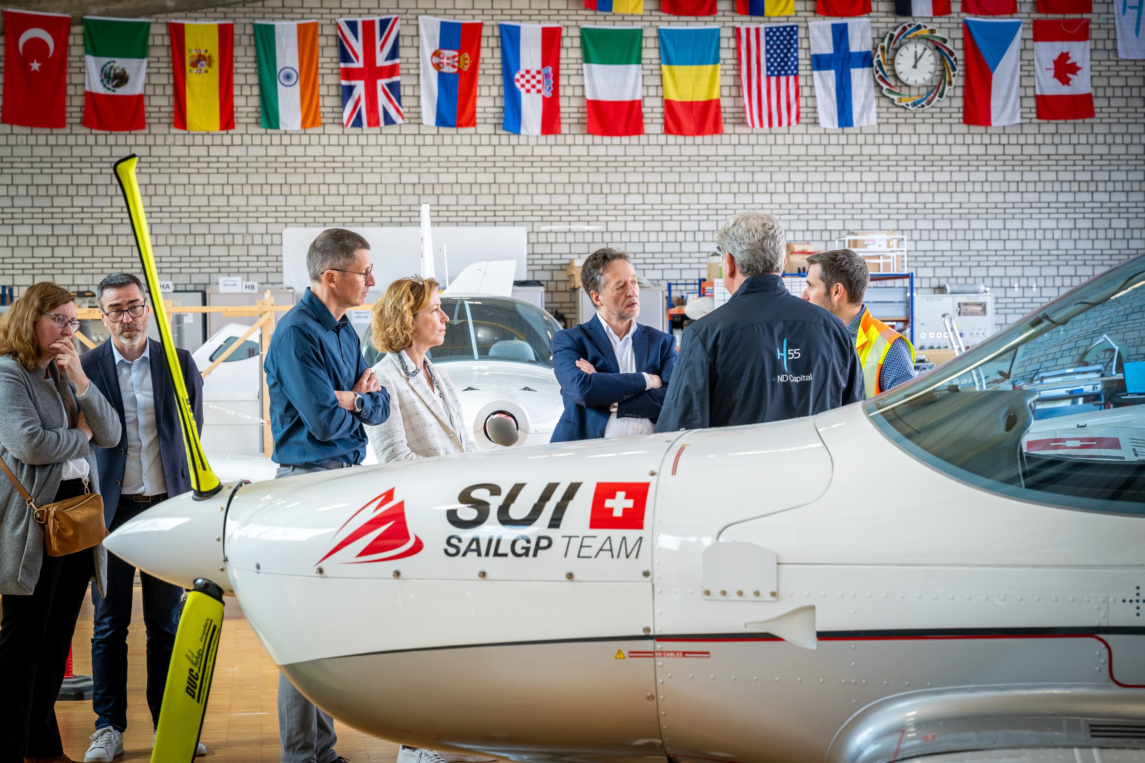 Délégation en visite devant le nez d’un avion électrique blanc, drapeaux du monde au mur du hangar