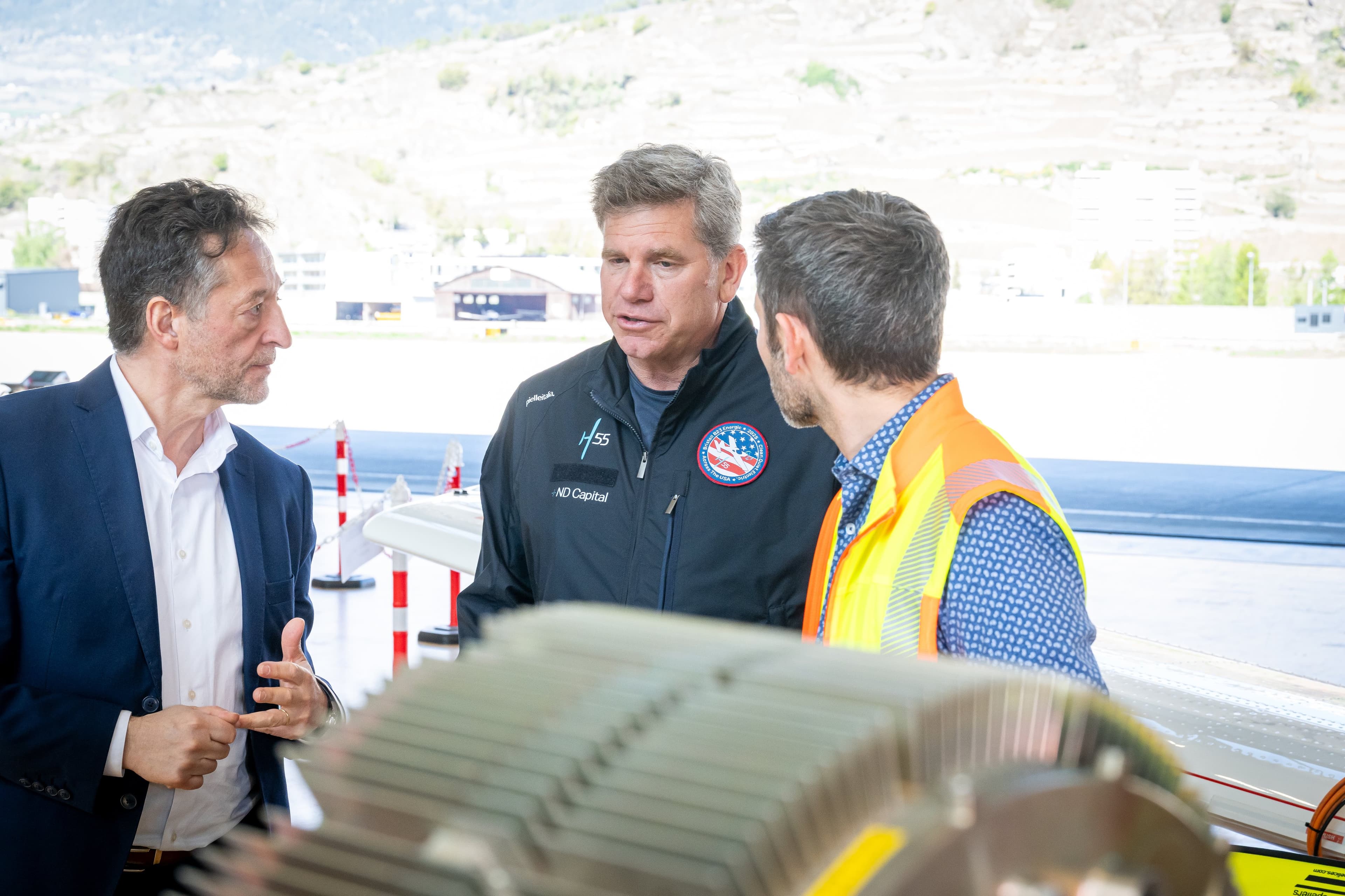 Trois hommes en discussion près d’un moteur électrique, piste et vignes en arrière-plan