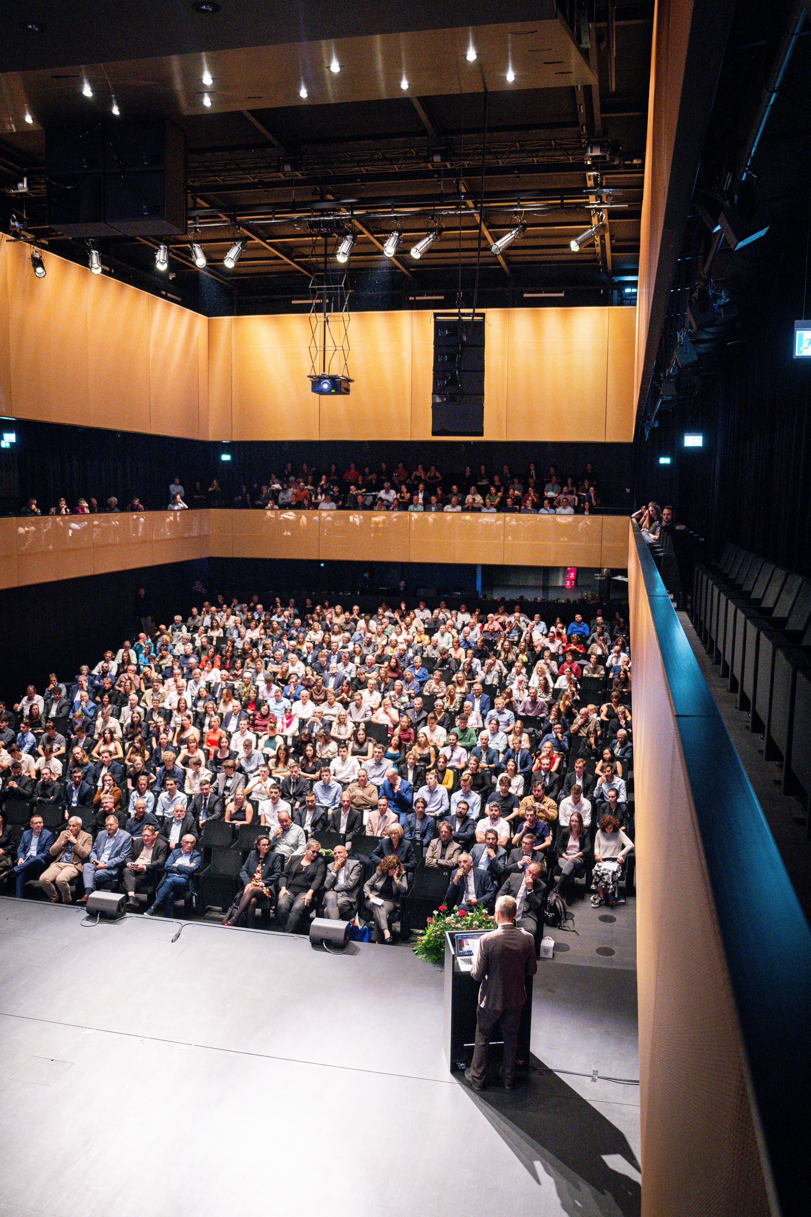 La salle comble vue depuis la scène, un orateur au pupitre