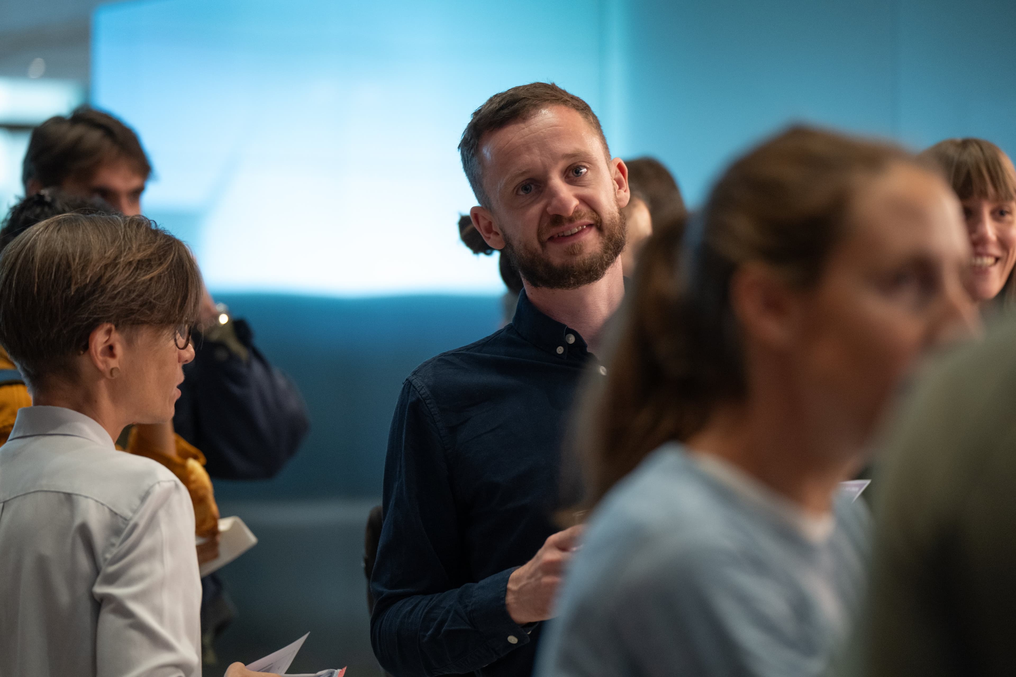 Un homme barbu écoute en souriant au milieu d’un groupe de participants, lumière bleue