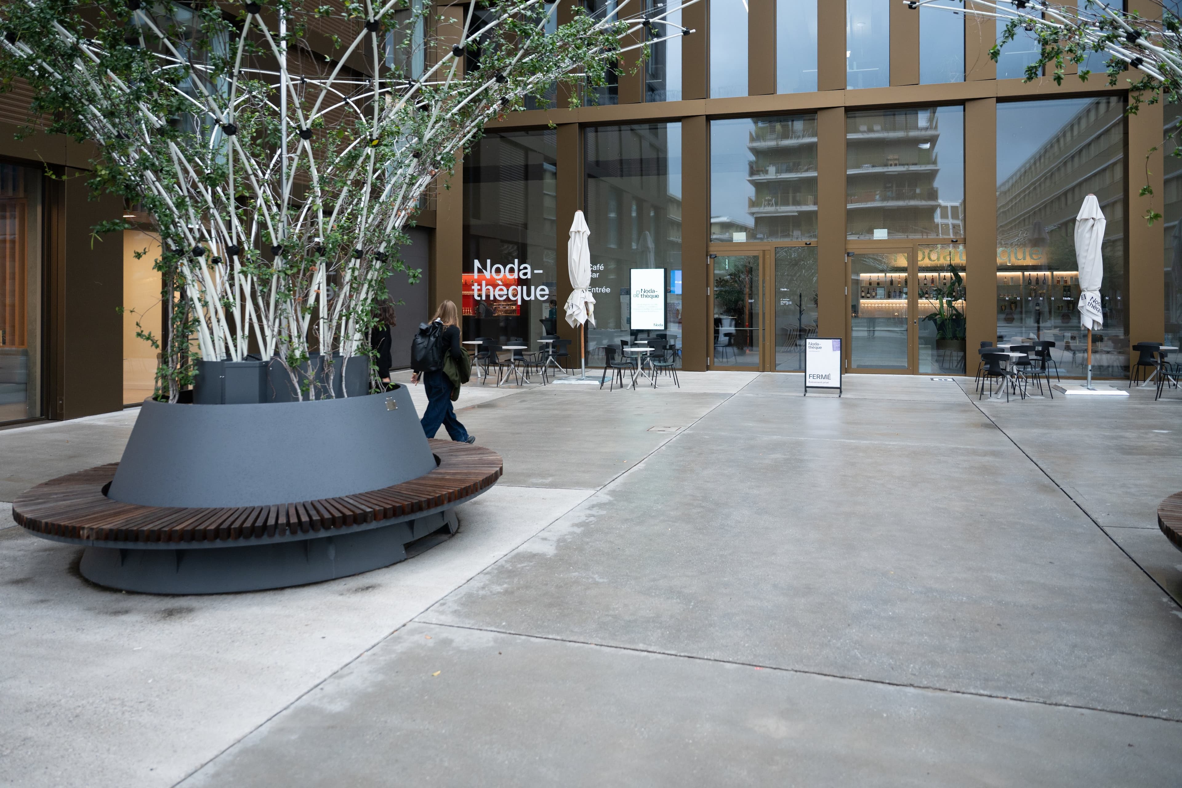 Parvis du Noda à Sion, un grand banc rond planté d’arbres devant la façade vitrée